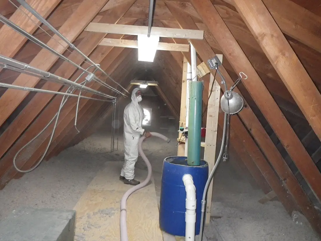 Worker in a protective suit blowing loose-fill insulation across an attic floor under wooden rafters