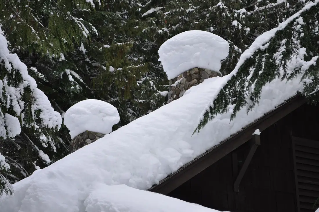 Deep snow piled on a sloped roof and two stone chimneys among snowy evergreen trees