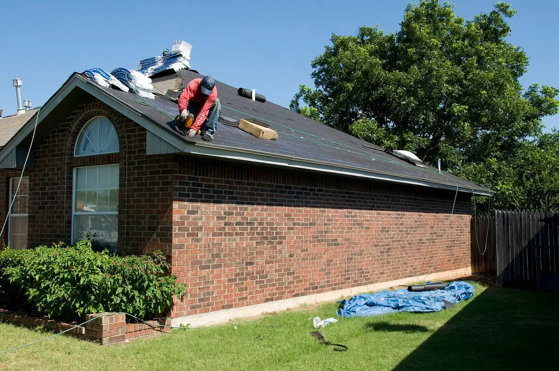 Roofer nailing shingles onto underlayment on the roof of a brick ranch house, with shingle bundles stacked near the ridge